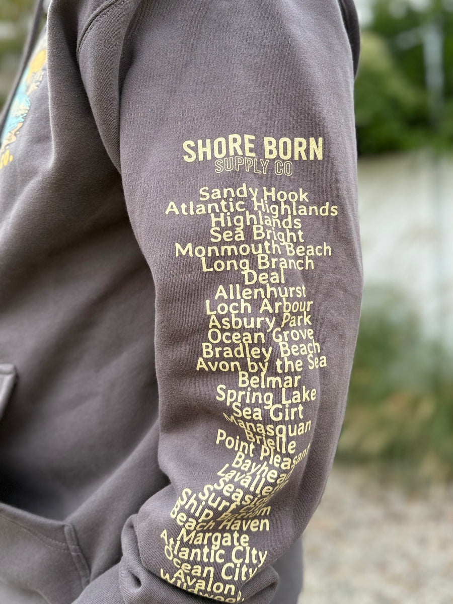 Gray sweatshirt with yellow text listing coastal towns on a blurred natural background
