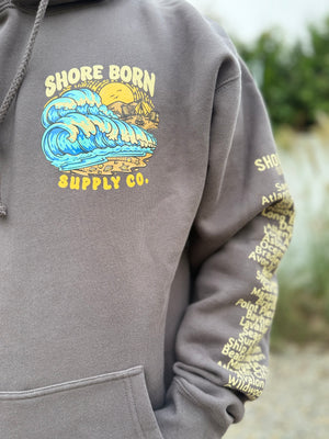 Gray hoodie with 'Shore Born Supply Co.' graphic on a blurred beach background