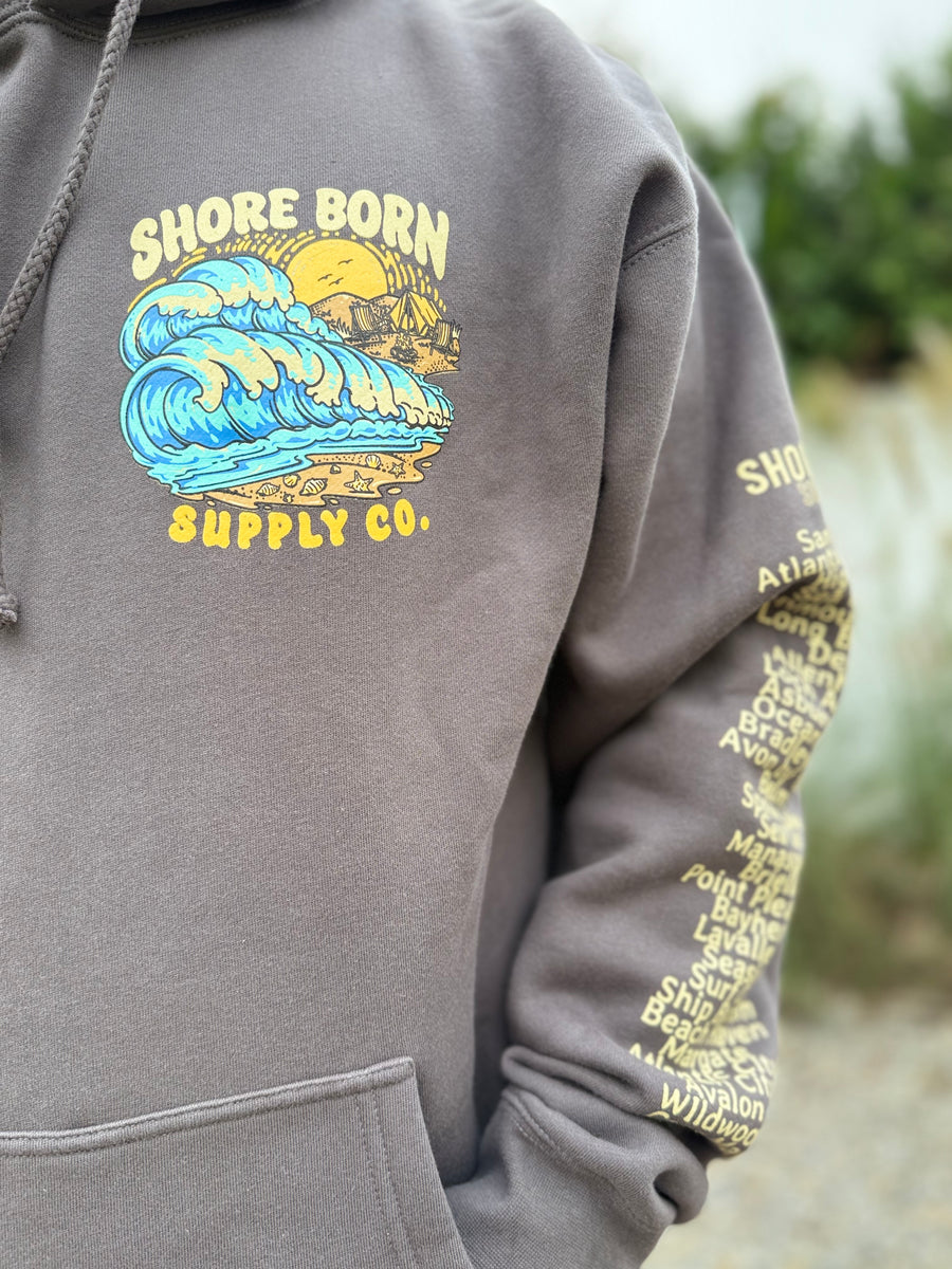 Gray hoodie with 'Shore Born Supply Co.' graphic on a blurred beach background